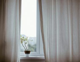 Foto de uma janela com cortinas brancas e um vasinho de flor na soleira.