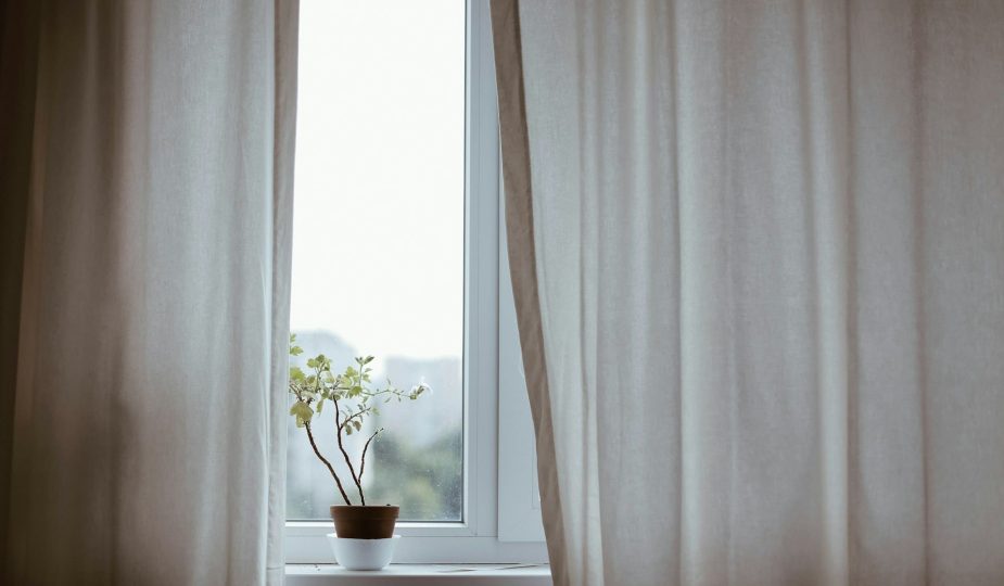 Foto de uma janela com cortinas brancas e um vasinho de flor na soleira.