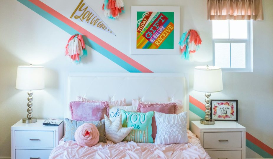 Foto de um quarto de adolescente com as paredes brancas, cama com colcha rosa e quadros na parede.