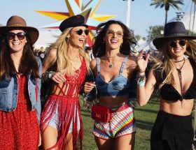 Foto com quatro mulheres com roupas de festival, óculos escuros e chapéu em um show.