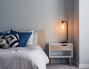 Foto de um quarto com paredes e cama branca, e uma mesinha com luminária.