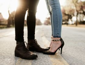 Foto das pernas de um casal vestindo calça jeans e sapatos, ele com bota e ela com salto alto.