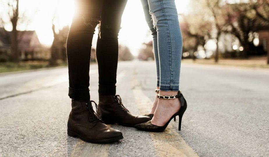 Foto das pernas de um casal vestindo calça jeans e sapatos, ele com bota e ela com salto alto.